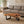 Load image into Gallery viewer, Ein massiver Freiform-Couchtisch aus Akazienholz steht vor einem taupefarbenen Sofa auf einem hellen Teppich. Die warme, lebhafte Holzmaserung, sichtbaren Jahresringe und natürlich geschwungene Baumkante machen die Tischplatte zum Blickfang. Schwarze Metallbeine bilden einen modernen Kontrast zum Holz. Seitliches Tageslicht, eine Zimmerpflanze und dezente Naturtöne verleihen dem Wohnzimmer eine helle, ruhige Atmosphäre.
