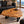 Laden Sie das Bild in den Galerie-Viewer, Holzstamm-Couchtisch aus massiver Suar-Baumscheibe (90 × 68 × 43 cm, 8 cm starke Platte) mit organischer Naturform, markanter Baumkante und sichtbarer Maserung, kombiniert mit schwarzem Ledersofa und warmem Massivholzboden – ein kompaktes Naturholz-Unikat für moderne, stilvolle Wohnzimmer in der Schweiz.
